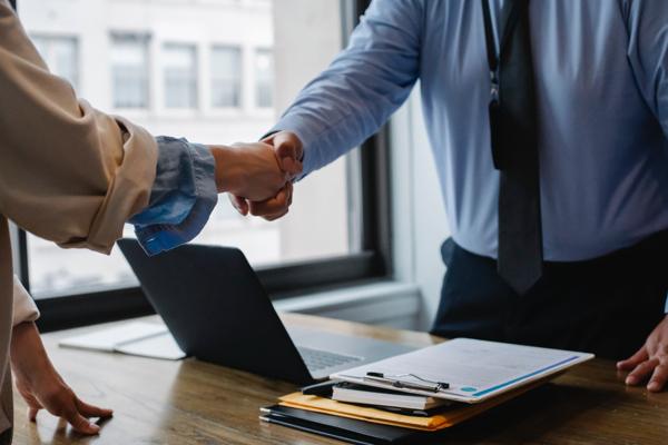 Two people in business attire shaking hands over a desk with a laptop and documents, near a large window.