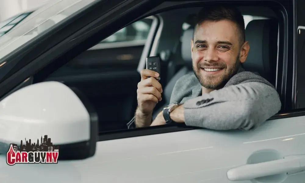 Smiling man in a car holding keys, wearing a gray blazer. CarGuvNY logo visible on side mirror.