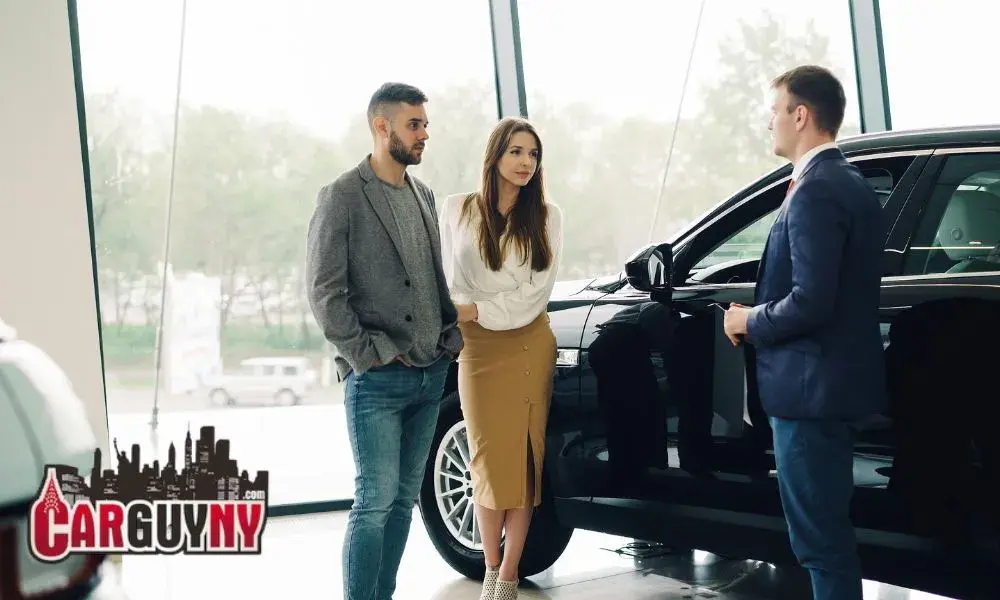A couple converses with a salesman next to a black car in a showroom, with large windows in the background and a "Car Guy NY" logo visible.