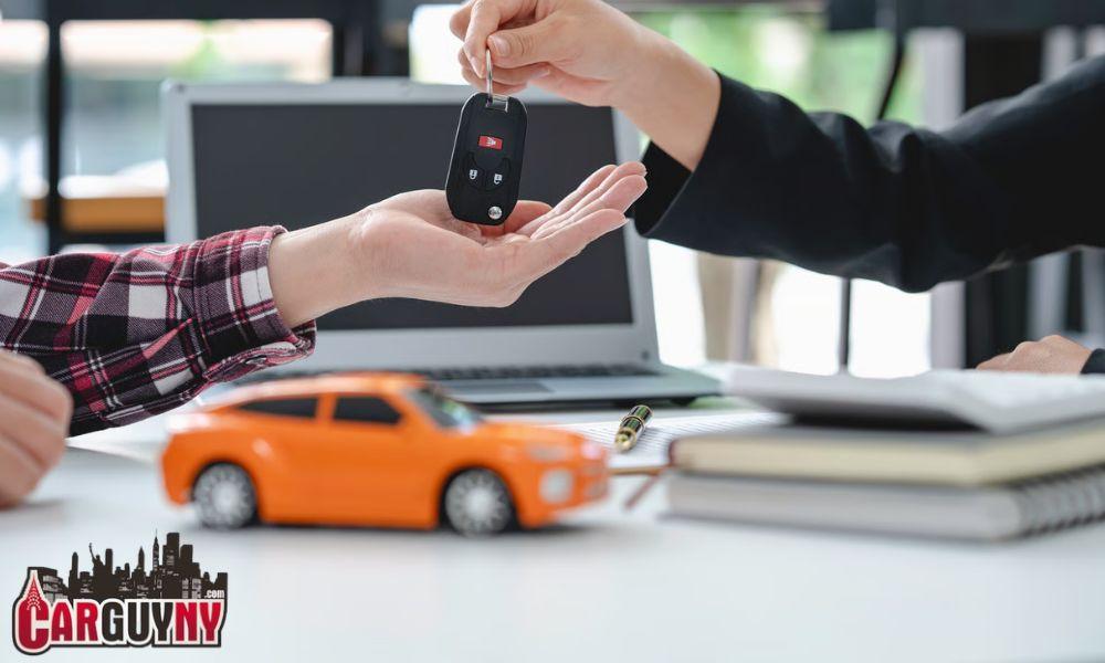 Handing over car keys above a table with a laptop, a small toy orange car, and some paperwork.