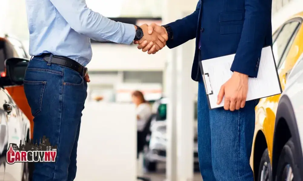 Two people shaking hands in a car dealership; one holds a clipboard, surrounded by cars.