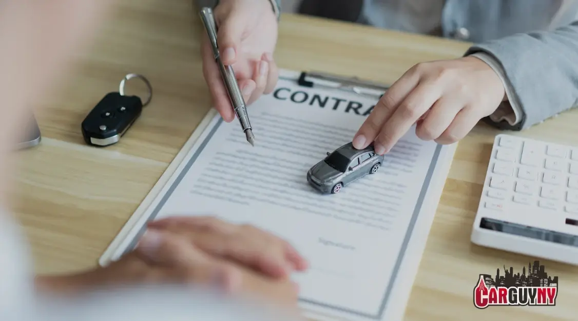 Hands signing a car contract with a small car model on the document. Car keys and a calculator are on the wooden table.