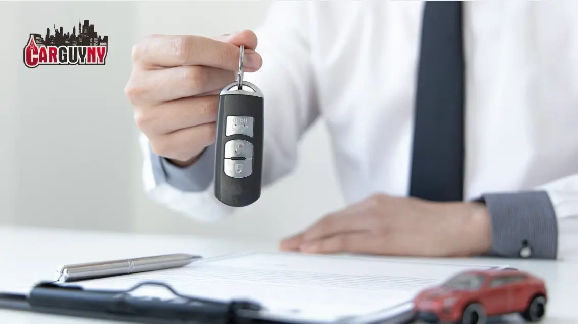 Man holding car key over signed contract with a toy car and pen on the table; "CarGuyNY" logo in the background.