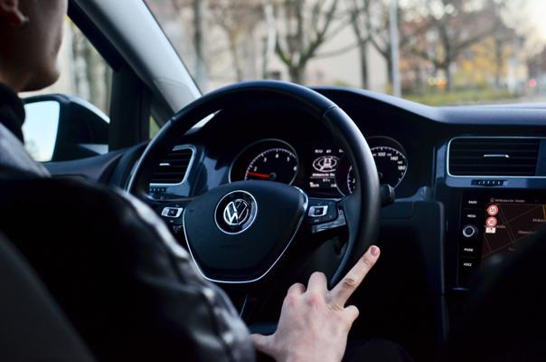 Person driving a Volkswagen car, hand on steering wheel, dashboard displaying speedometer and gauges, trees visible through windows.
